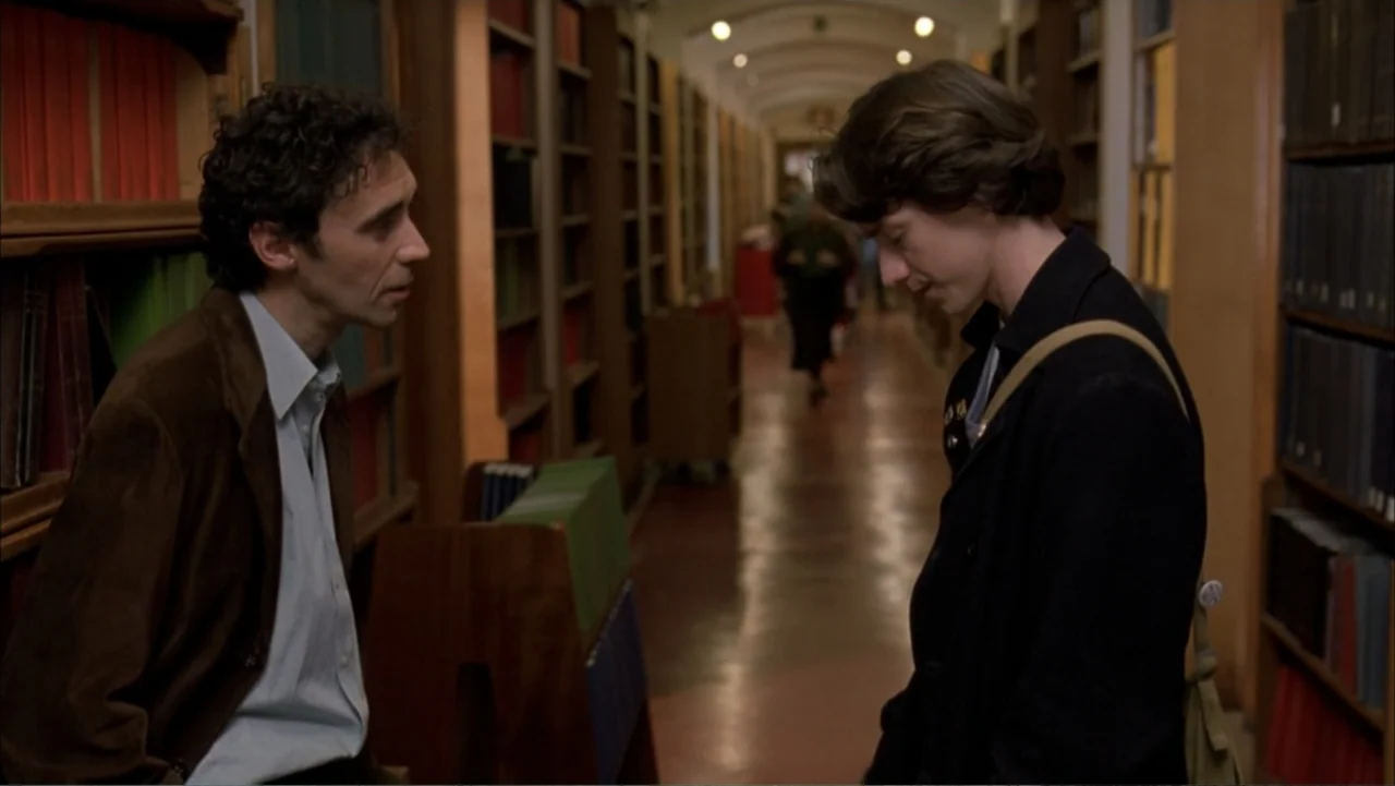 Starter for 10 (2006) movie still: medium — A long aisle of a library is filled with towering rows of books. Two men stand facing each other while…