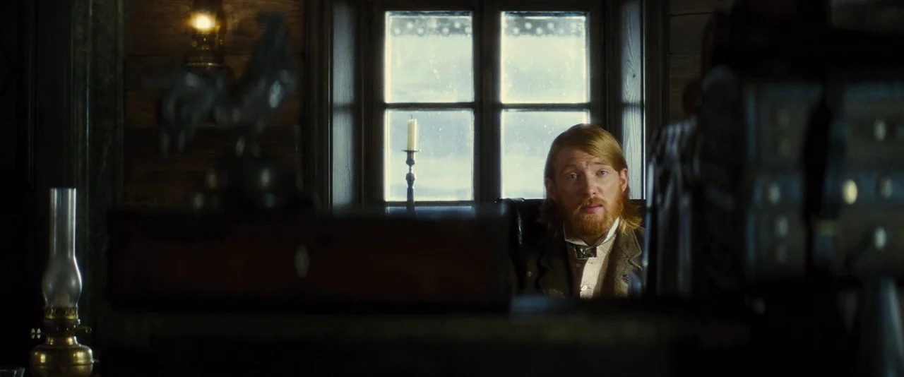 Anna Karenina (2012) movie still: medium — A dark, wood-paneled room is lit by a large window and interior candles. A man sits at a desk in the…