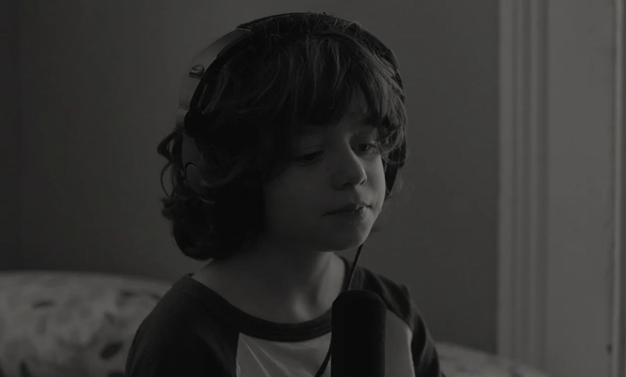 C’mon C’mon (2021) movie still: closeup — A close-up shot captures a young boy wearing large over-ear headphones. He stands before a microphone,…