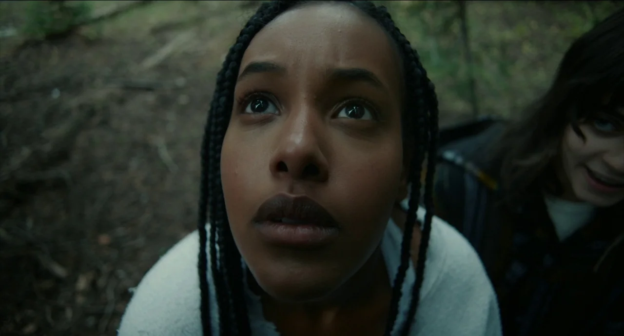 Trim Season (2023) movie still: closeup — A dense, leaf-covered woodland floor serves as the background for two people. A young woman with braids…