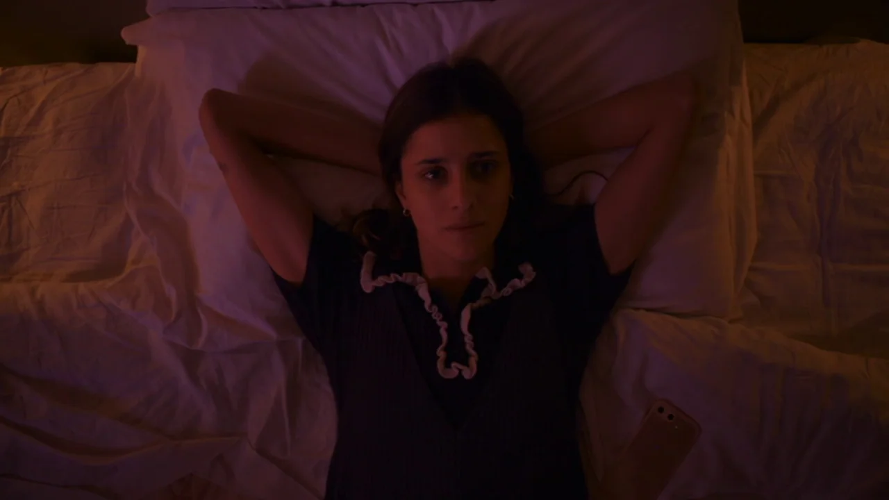 Amanda (2022) movie still: medium — A woman lies flat on a white pillow covered bed in a dark room. She rests with her hands behind her head while…