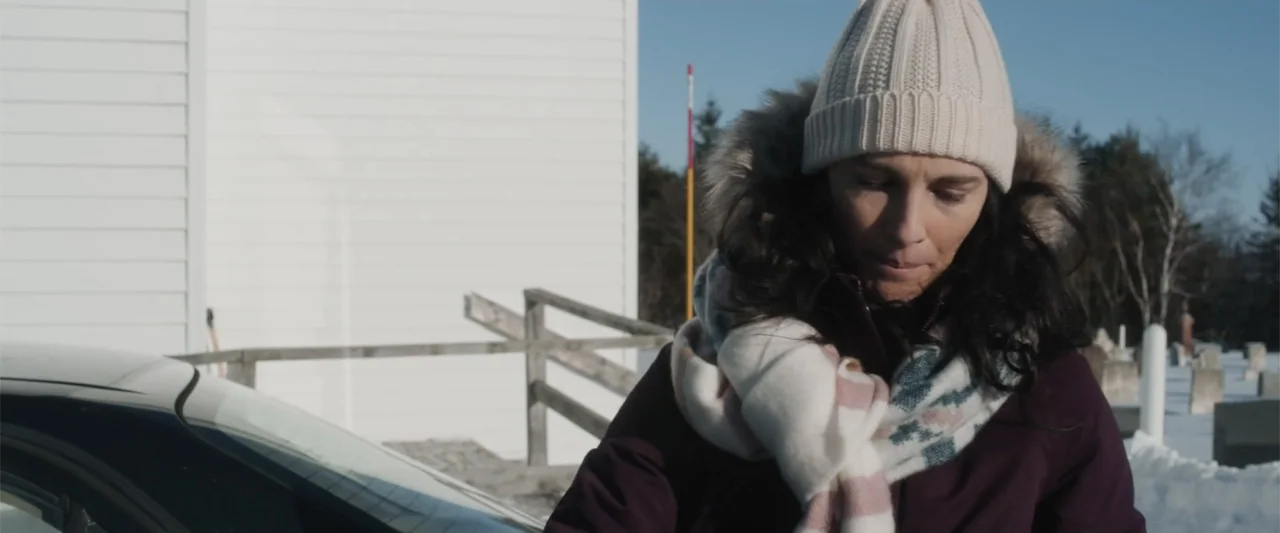 A Small Fortune (2021) movie still: medium — A woman stands in a snowy cemetery with a white building and fence nearby. She wears a winter coat and…