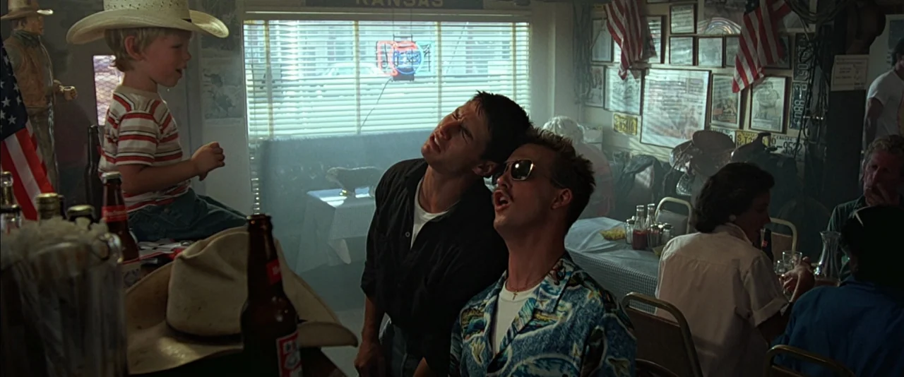 Top Gun (1986) movie still: medium — An interior shot showing a wood-paneled establishment decorated with American flags and framed photographs. Two…