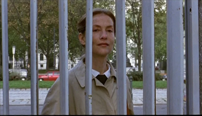 The Piano Teacher (2001) movie still: medium — A woman stands behind a series of vertical metal bars in an outdoor setting. The background includes…