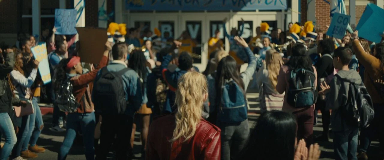 Freaky (2020) movie still: wide — A crowd of students gathers in front of a brick high school building exterior. They hold handmade signs and wear…