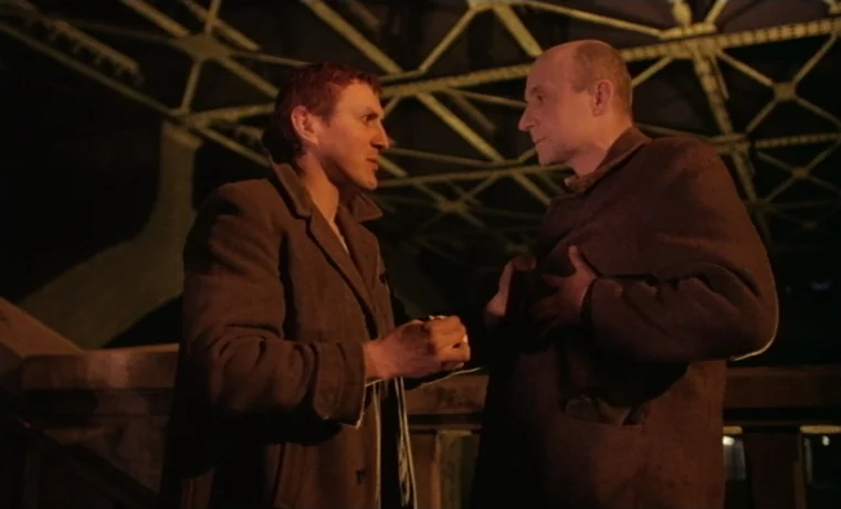 Golem (1980) movie still: medium — Two men stand under an industrial steel bridge structure at night. They face each other in conversation while…