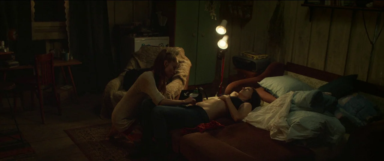 Nightsiren (2022) movie still: wide — A dimly lit room with wood-paneled walls contains a bed and sparse furniture. One woman kneels on the bed…