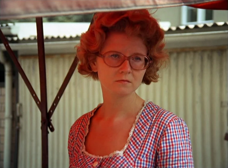 The Merchant Of The Four Seasons (1971) movie still: closeup — A woman stands outdoors under the edge of a red market stall canopy. She looks past…