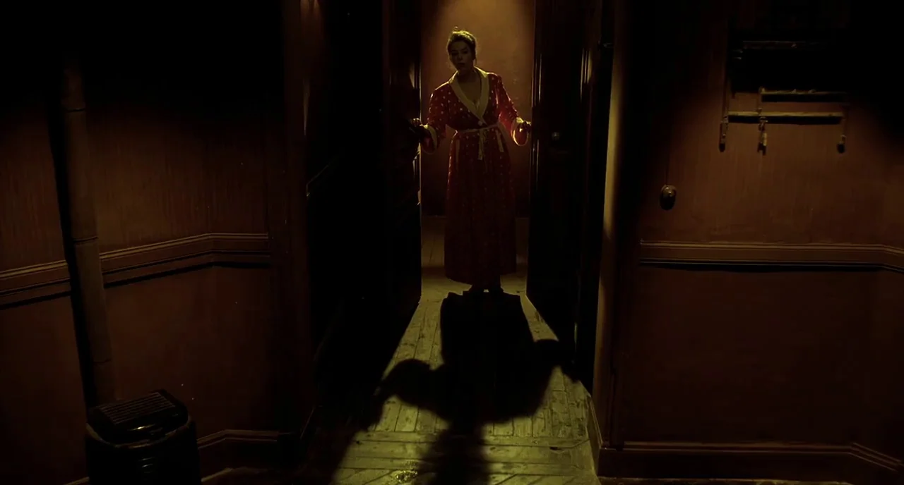 Delicatessen (1991) movie still: fullbody — A dimly lit narrow corridor features dark walls and a worn wooden floor. A woman in a red patterned…