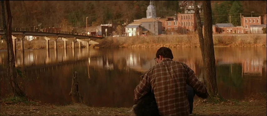 All The Real Girls (2003) movie still: medium — A man sits by a calm river with a small town visible across the water. The man faces away from the…