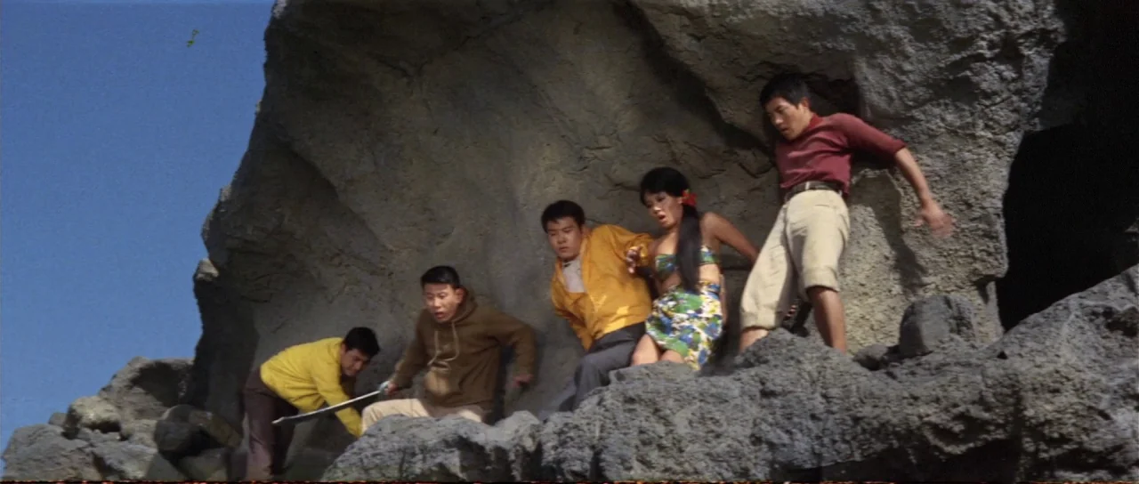 Ebirah Horror of the Deep (1966) movie still: fullbody — A group of five people traverses a rough, dark volcanic rock formation outdoors. The…
