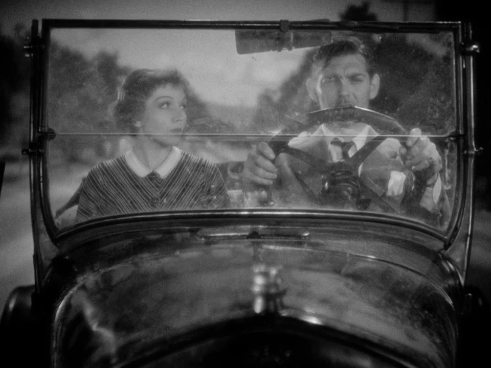 It Happened One Night (1934) movie still: medium — A man and a woman sit inside a vintage automobile at night. The man operates the steering wheel…