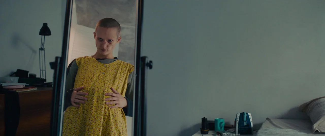 Titane (2021) movie still: medium — A plain room with a stand-alone mirror reflecting a person in a floral dress. The individual stands in front of…