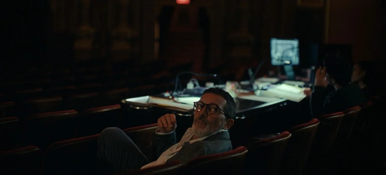 Babygirl (2024) movie still: medium — A dark, empty theater auditorium contains rows of red velvet seats and a technical console. A man sits in the…