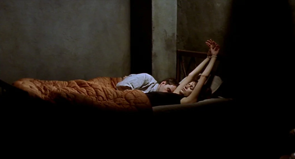 Tiresia (2003) movie still: medium — A stark room with grey concrete walls contains a simple bed frame. A man lies next to a woman whose wrists are…