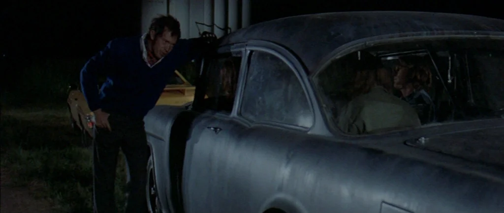 Two Lane Blacktop (1971) movie still: medium — A man stands outdoors at night next to a dark, worn-in vintage sedan. He leans against the driver…
