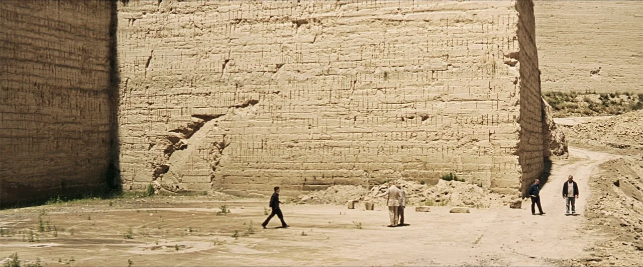 GOMORRAH (2023) movie still: wide — A vast limestone quarry features towering vertical rock faces and a dusty floor. Four men stand in a group at…