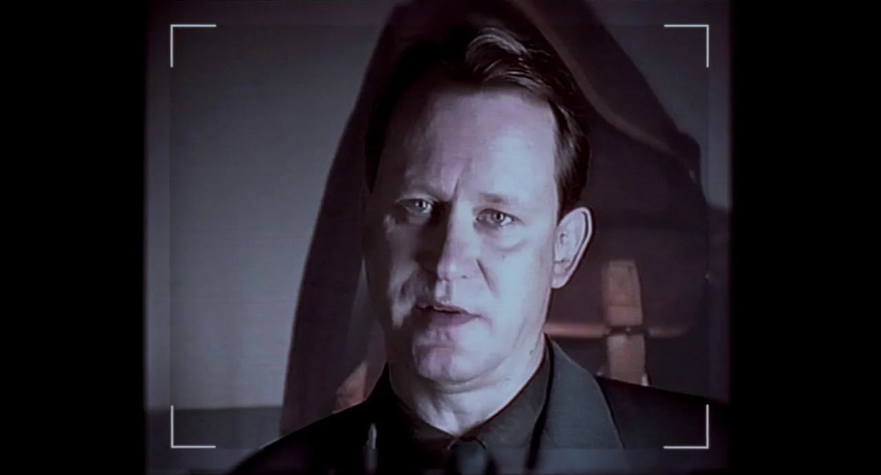 Insomnia (1997) movie still: closeup — A man is centered in a dimly lit space with a blurred dark backdrop. He is framed within a digital viewfinder…