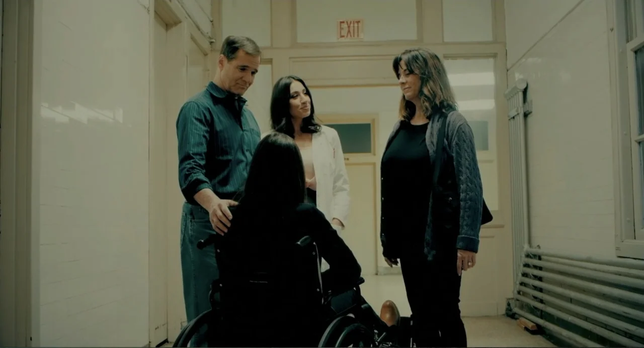 Terrifier (2018) movie still: fullbody — A hospital hallway features white tiled walls and a bright exit sign overhead. Three people stand around a…