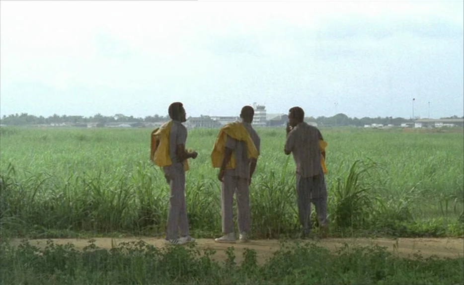 Chocolat (1988) movie still: fullbody — Three men stand on a dirt path bordered by a large expanse of green grass. A small control tower is visible…