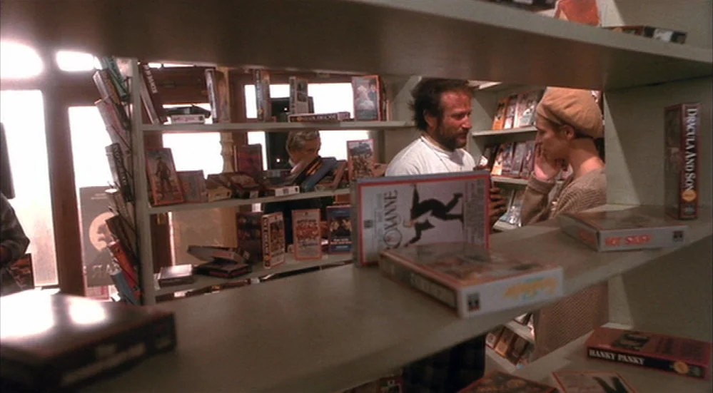 The Fisher King (1991) movie still: medium — A video rental store with shelves filled with VHS tapes in plastic cases. A man and a woman stand…