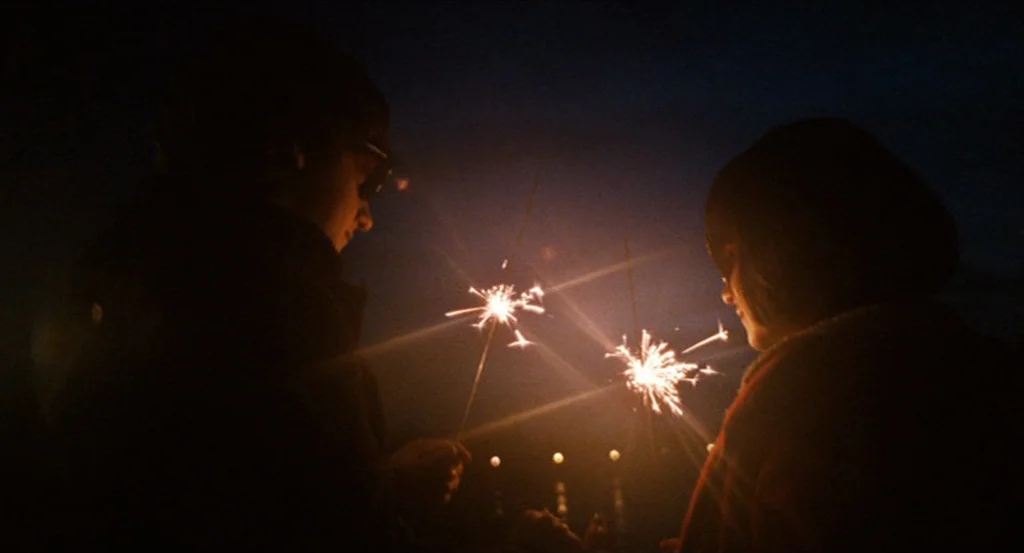 Submarine movie still: medium — Two individuals stand in total darkness outside at night holding lit sparklers. The bright flares illuminate their…