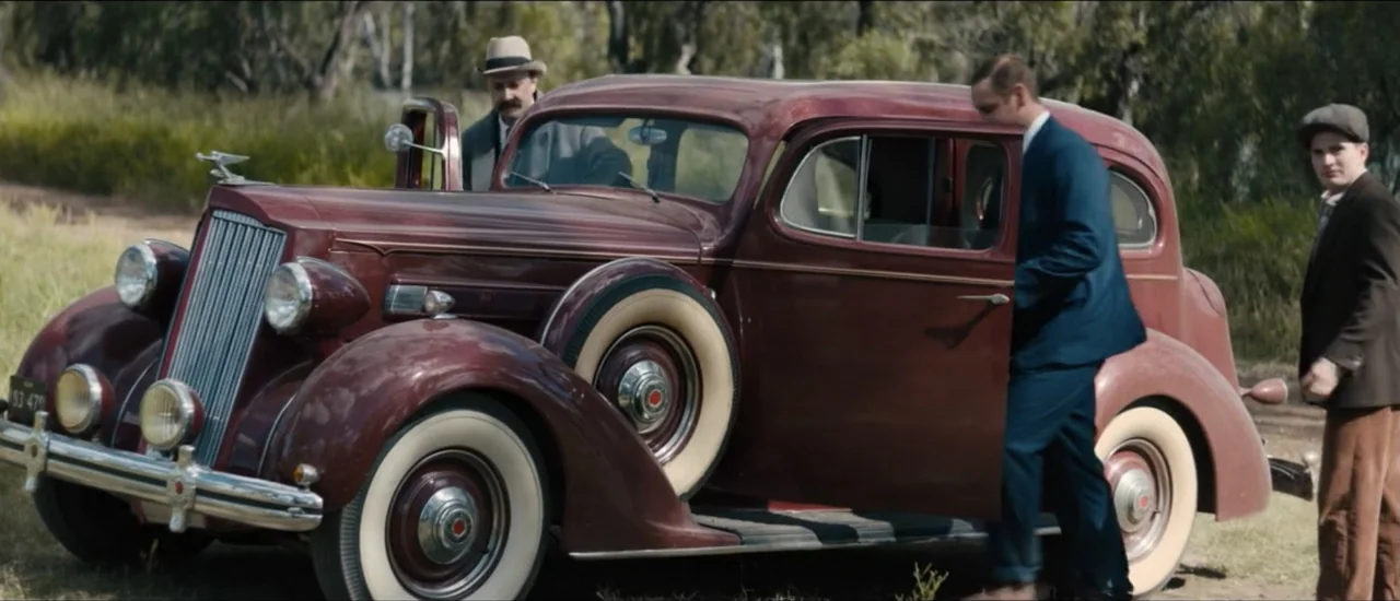 Fear Below (2025) movie still: wide — A dark red 1930s sedan is parked on a dusty rural road. Three men stand beside the vehicle in an open…