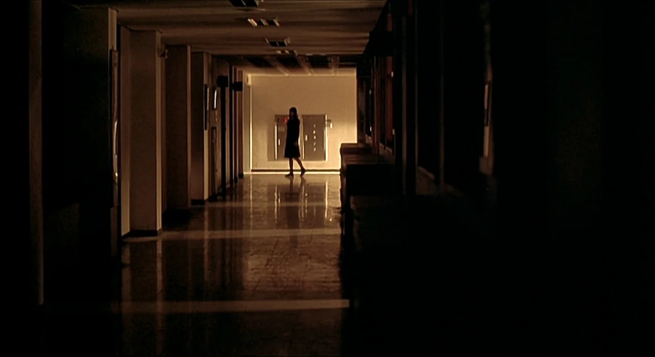 Voice (2005) movie still: wide — A long, dimly lit institutional hallway with a tiled, reflective floor surface. A single woman walks slowly across…
