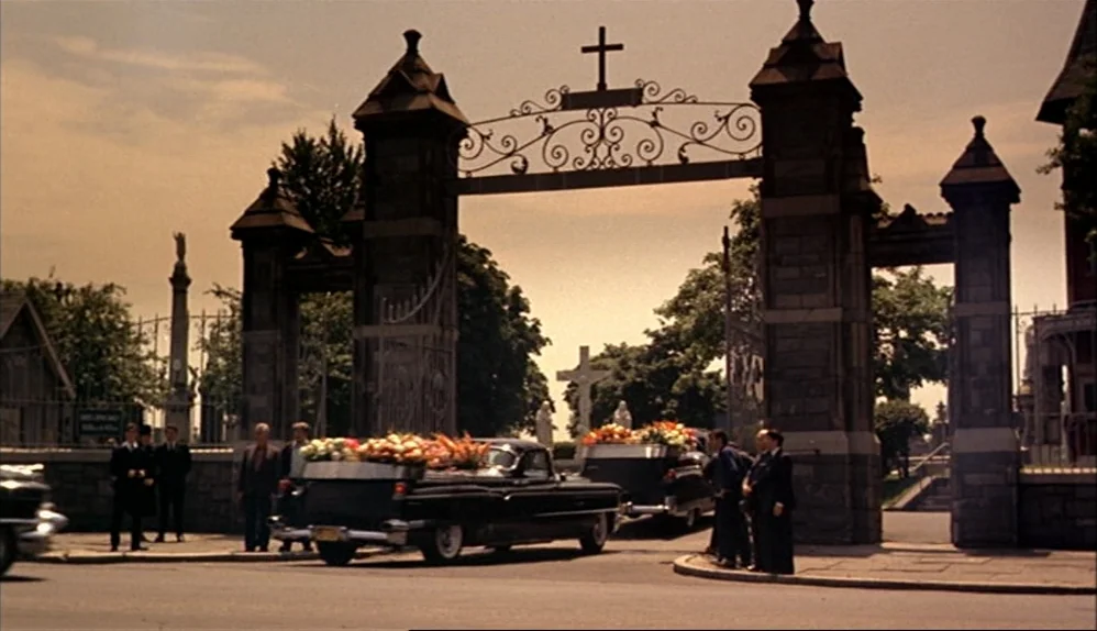 The Godfather (1972) movie still: wide — A stone cemetery gate with a cross sits behind two black vintage funeral cars. Several men in suits stand…