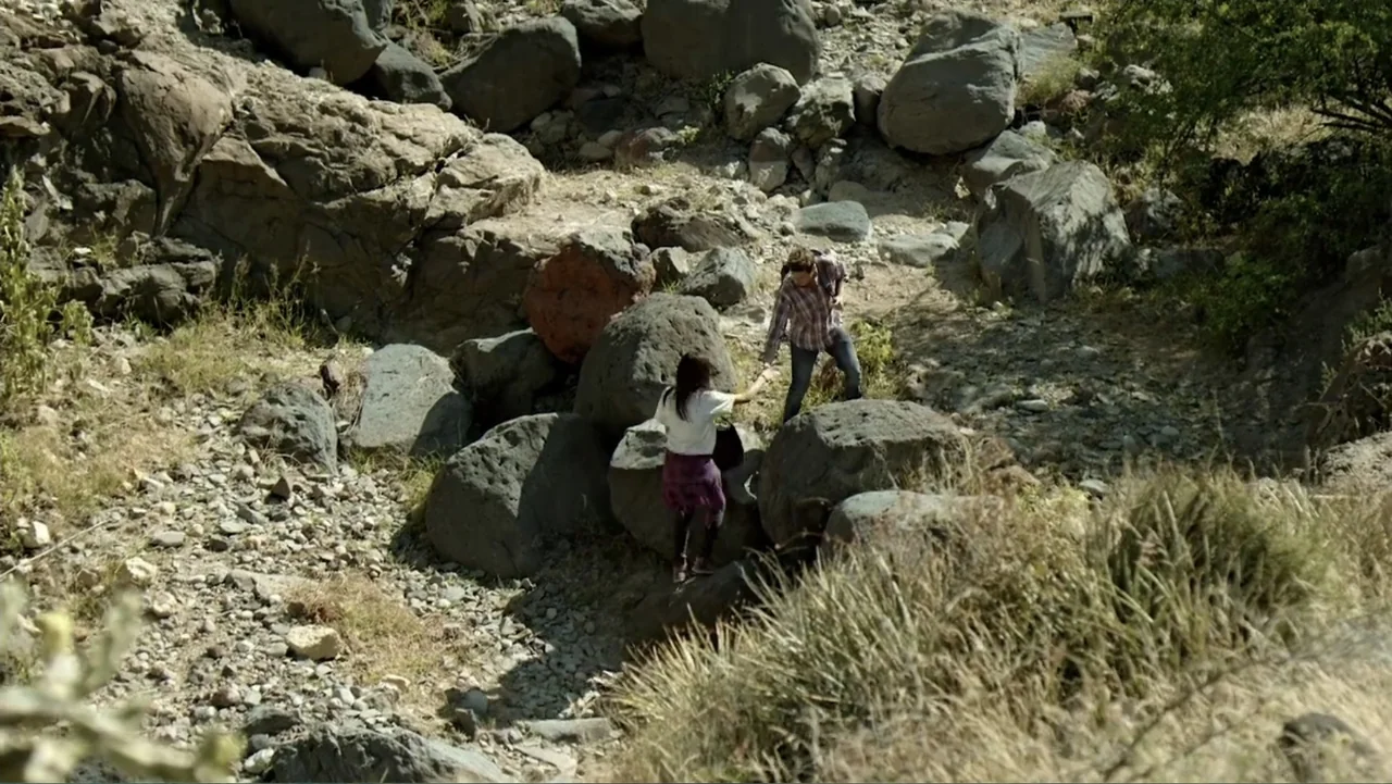 Loco Love (2003) movie still: wide — A dry, rocky ravine is filled with large boulders and sparse, dry vegetation. A woman helps a man climb over a…