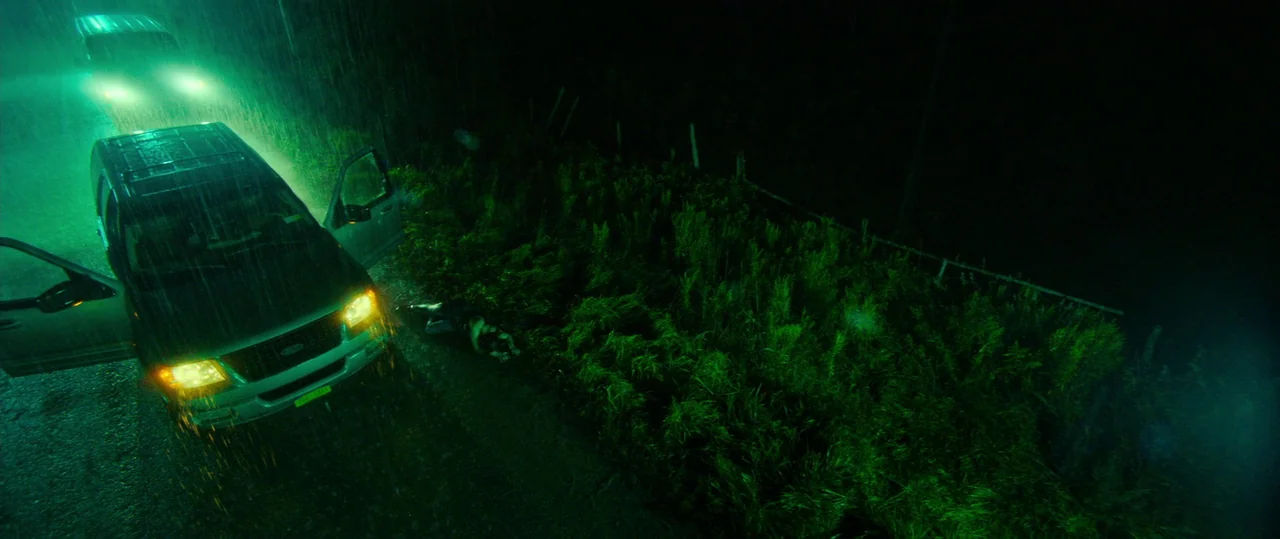 Random Acts of Violence (2019) movie still: wide — A dark, rain-slicked roadside area features tall grass and a parked SUV at night. A person lies…
