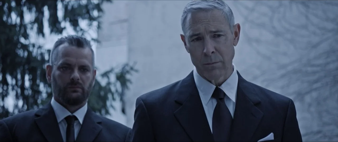 Room 6 (2006) movie still: medium — An outdoor location features two men in formal attire standing in front of trees. The men remain still as they…