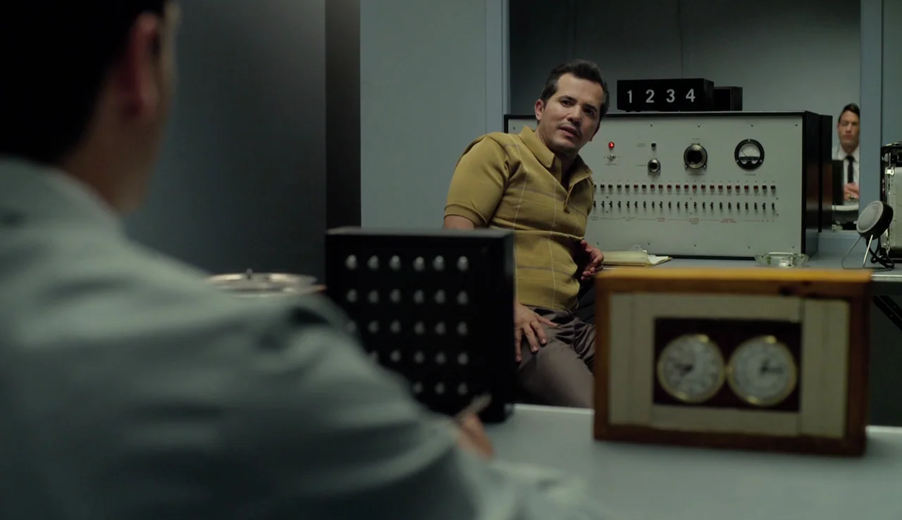 Experimenter (2015) movie still: medium — A man sits across from another person in a sterile laboratory environment. The room features a large…