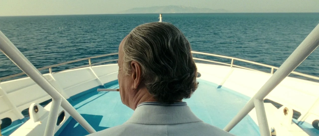 The Great Beauty (2013) movie still: medium — The bow of a white and blue boat sits on the open ocean under daylight. An elderly man in a white suit…