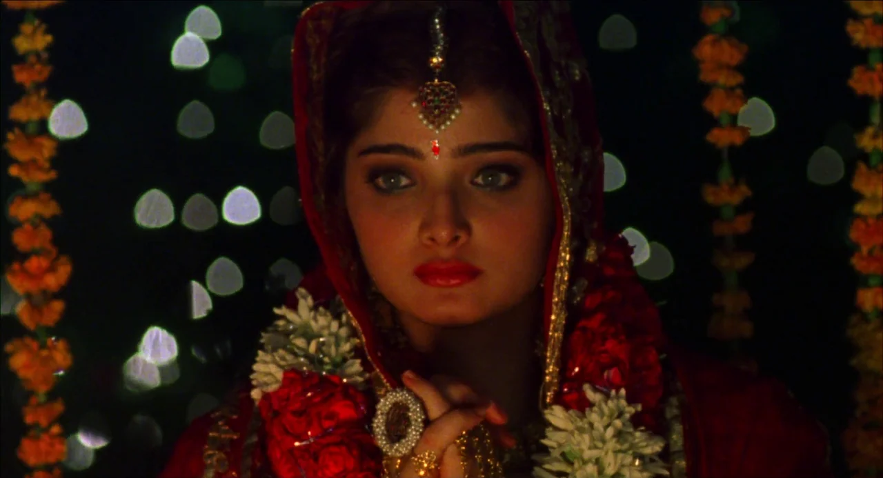 Monsoon Wedding (2001) movie still: closeup — A dark, blurred background features hanging strings of orange marigold lights in the distance. A woman…