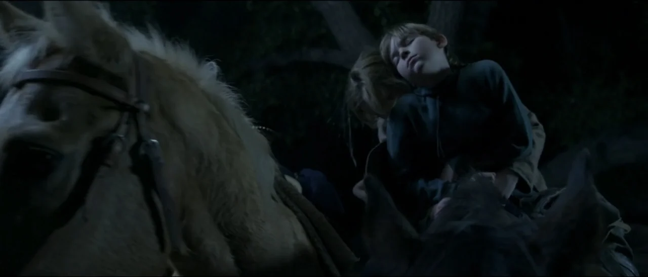 Dead for a Dollar (2022) movie still: medium — A dark outdoor space at night features dense foliage and deep shadows. Two children ride on the back…