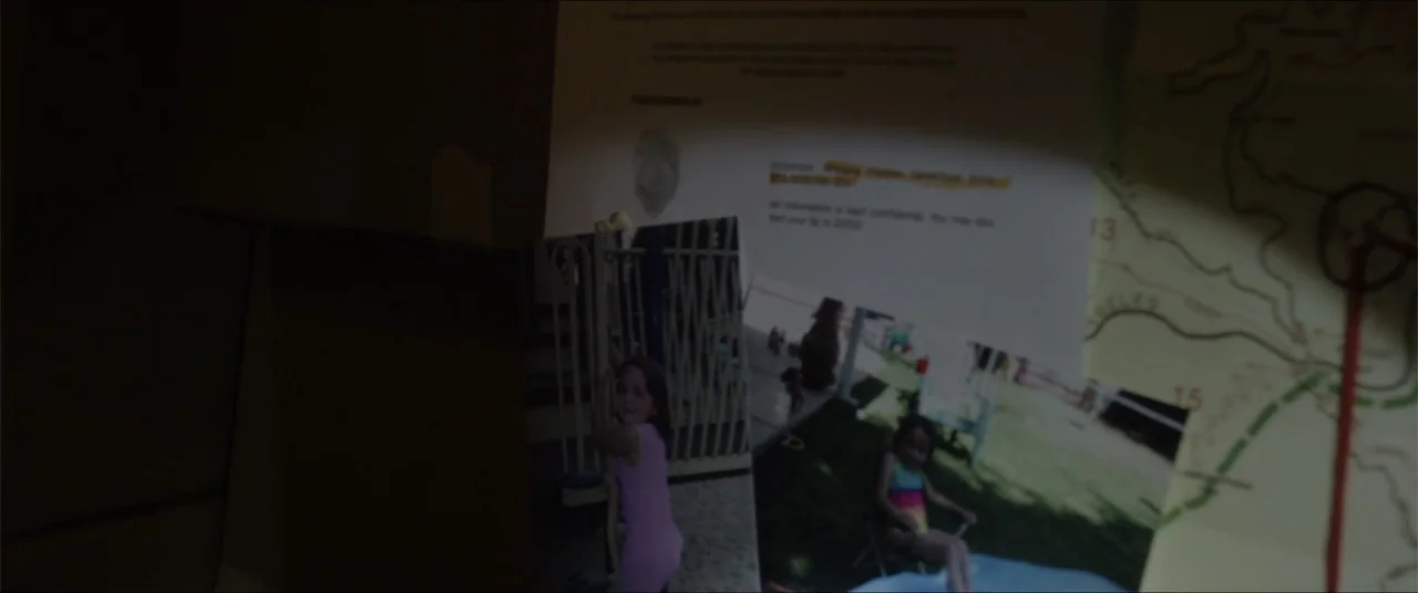 Captive (2020) movie still: closeup — A dark indoor wall surface features several pinned photographs of a young child. A bright light illuminates a…