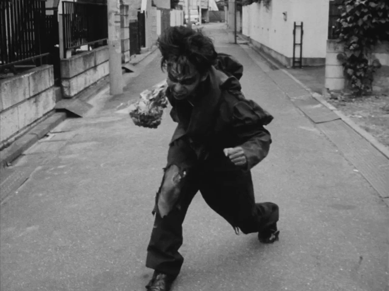 Tetsuo: The Iron Man (1989) movie still: fullbody — A narrow paved residential street with white walls and a black fence. A man in torn dark…