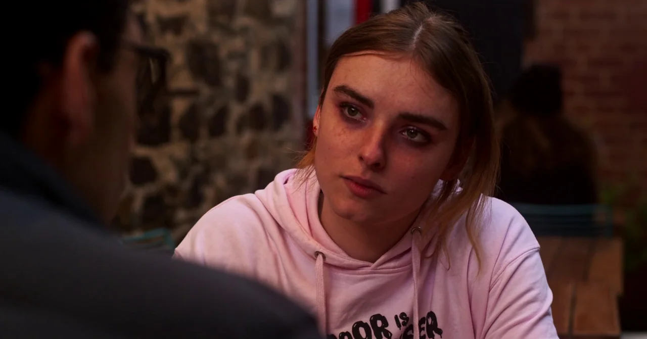 T Blockers (2023) movie still: medium — A woman sits at a wooden table in an indoor space with brick walls. She looks toward an out-of-focus person…