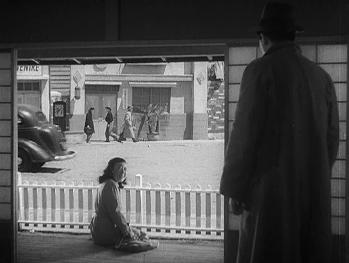 Anatomy of a Relationship (1976) movie still: fullbody — A wooden veranda overlooks a bright street scene with pedestrians and a parked car. A man…
