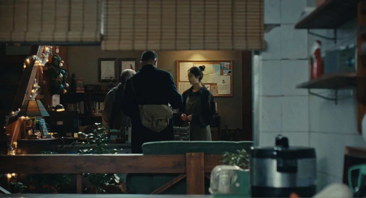 Winter in Sokcho (2025) movie still: medium — A cafe interior with dark wood furniture and shelves adorned with small holiday decorations. A man in…