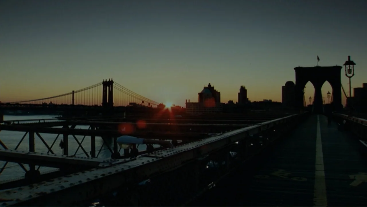 The Words (2012) movie still: establishing — A view across a metallic bridge walkway toward a distant suspension bridge at sunrise. The city skyline…