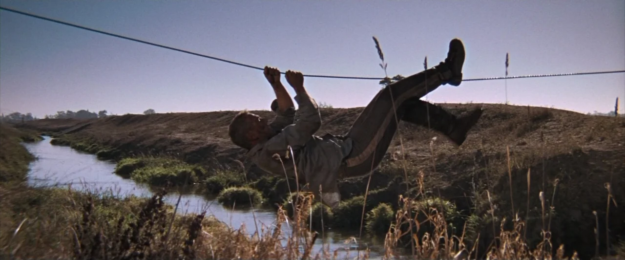 Cool Hand Luke (1967) movie still: wide — A man slides along a wire over a narrow creek in a barren landscape. The area features dry, patchy grass…