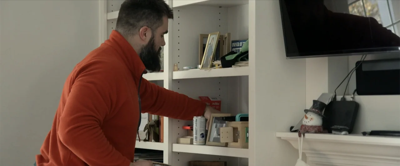 Kelce (2023) movie still: medium — A white built-in bookshelf sits against a wall containing various framed photos and objects. A bearded man in an…