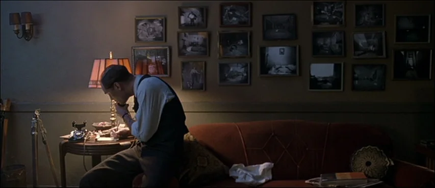 Road To Perdition (2002) movie still: medium — A dimly lit room features a red sofa and walls covered with many framed photographs. A man sits on…