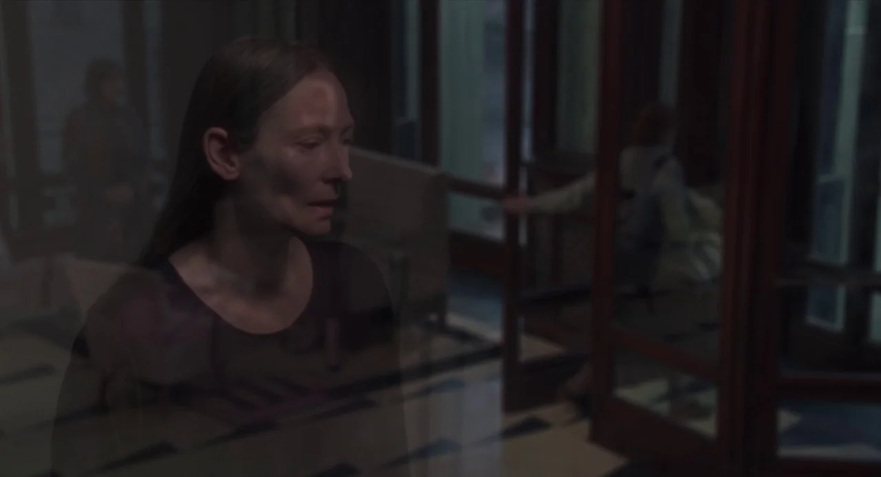 Suspiria (2018) movie still: medium — A modern building lobby seen through a glass pane with a reflection on the surface. A woman stands in the…