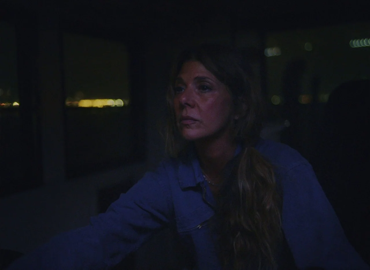 She Came To Me (2023) movie still: medium — A dimly lit interior space at night overlooks city lights through a dark window. A woman with long wavy…