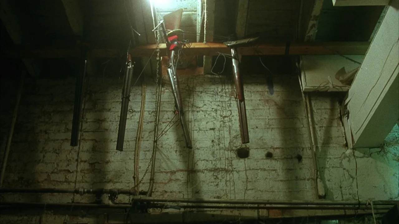 Saw movie still: wide — A rough, unfinished basement features white painted brick walls and exposed wooden ceiling beams. Several hunting rifles…