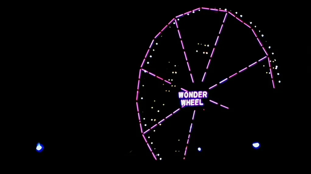 The Warriors (1979) movie still: establishing — A large Ferris wheel stands illuminated against a pitch-black night background. The structure…