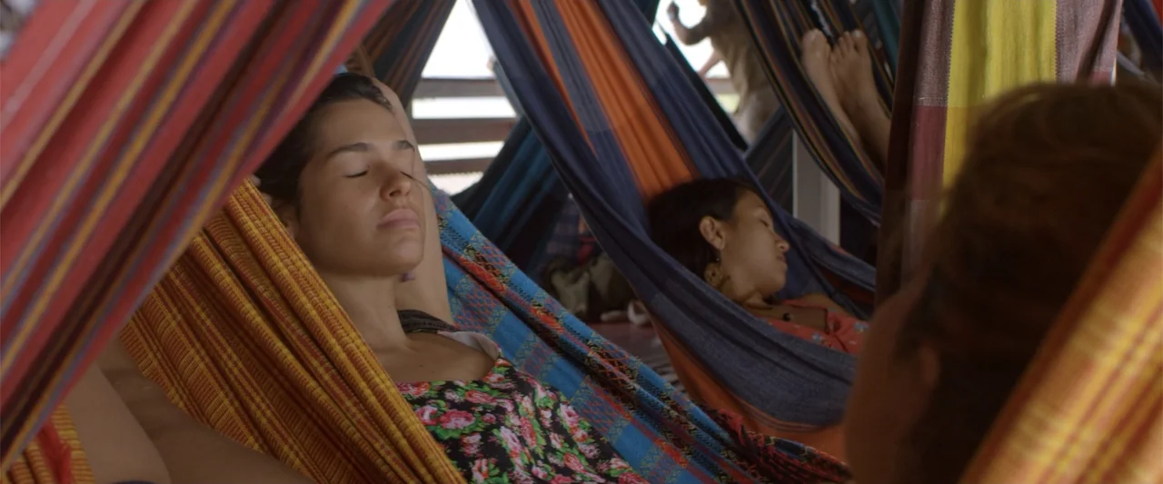 Dirty Little Secret (1998) movie still: medium — A group of multi-colored woven fabric hammocks hangs under a covered structure outdoors. A woman…